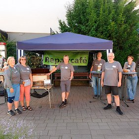 Gruppe von Personen an einem Infostand mit blauem Pavillon und "Repair Café"-Banner im Freien
